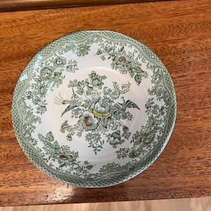 May include: A round ceramic bowl featuring a cream-colored base with a detailed green transferware pattern. The design includes intricate floral arrangements, foliage, and two bird figures. The rim has a textured, cross-hatched green border.
