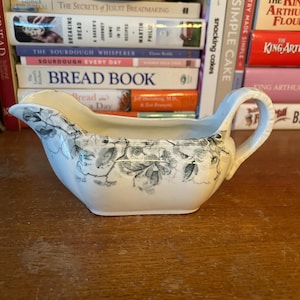 May include: A white and gray floral patterned gravy boat with a handle. The boat is made of ceramic and sits on a wooden surface. The background includes cookbooks and a white bowl.