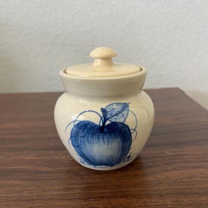 May include: A small, cream-colored ceramic jar with a lid. The jar is decorated with a blue apple design. The apple is detailed with a stem and leaf. The jar is sitting on a dark wooden surface.