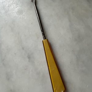 May include: A vintage crochet hook with a curved metal hook and a yellow, geometric handle. The hook is made of metal and has a dark finish. The handle is a bright yellow color.