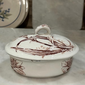 May include: A white ceramic serving dish with a lid, featuring a brown floral and botanical design. The dish has a rounded shape with a small handle on the lid. The design includes flowers and stems, creating a vintage aesthetic.