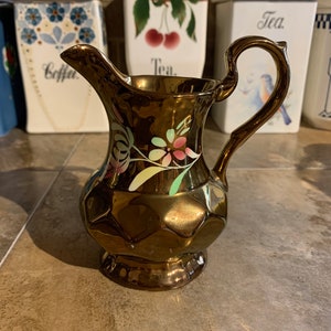 May include: A copper colored pitcher with a floral design in pink, green, and yellow. The pitcher has a textured surface and a handle.