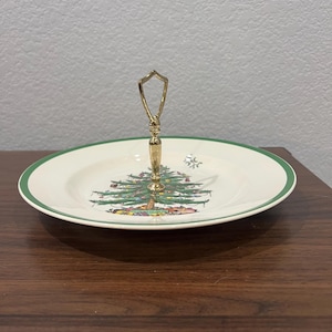 May include: A white ceramic serving tray with a green rim and a gold handle. The tray features a Christmas tree design with a green tree and colorful ornaments.