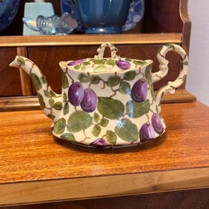 May include: A ceramic teapot with a lid, featuring a cream-colored base adorned with purple plums and green leaves. The teapot has a curved spout and handle, and is approximately 6 inches in height.