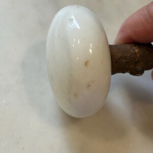 May include: A round, white porcelain door knob with a smooth, glossy finish and small brown surface imperfections. The knob is attached to a dark, textured, and rusted metal spindle that extends from the back of the white ceramic component.