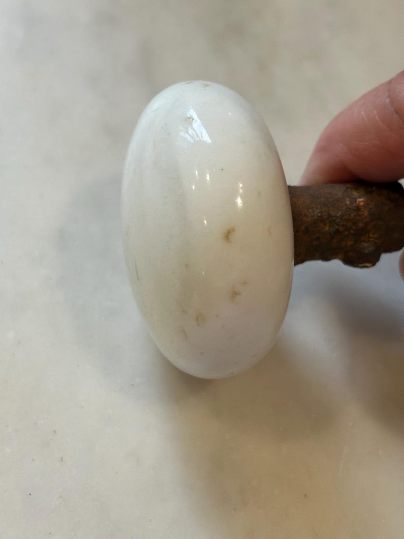 May include: A round, white porcelain door knob with a smooth, glossy finish and small brown surface imperfections. The knob is attached to a dark, textured, and rusted metal spindle that extends from the back of the white ceramic component.