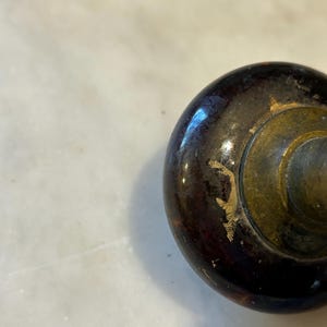 May include: Close-up of a dark brown, slightly worn wooden knob with a brass-colored metal base. The knob has a glossy finish with some areas of paint loss, revealing the wood underneath. The background is a light-colored surface.