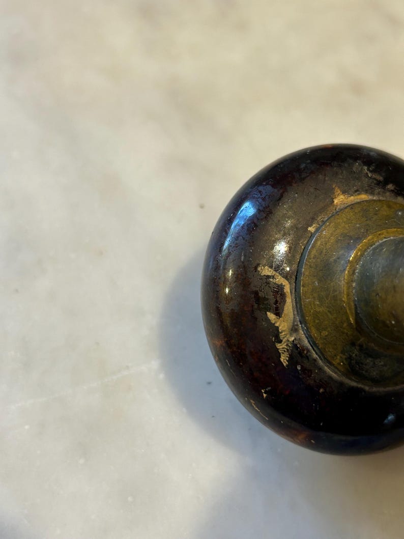May include: Close-up of a dark brown, slightly worn wooden knob with a brass-colored metal base. The knob has a glossy finish with some areas of paint loss, revealing the wood underneath. The background is a light-colored surface.