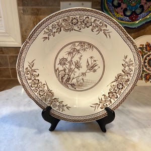 May include: A cream-colored ceramic plate featuring a brown transferware pattern. The center depicts a bird perched on bamboo, while the rim is decorated with floral motifs and a geometric border. The plate rests on a black plastic stand.
