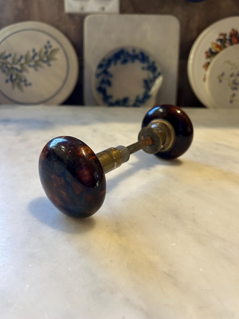 May include: Antique door knob with a dark brown, marbled finish. The knob has a rounded shape and is attached to a brass stem. The background features decorative plates with floral and leaf patterns.