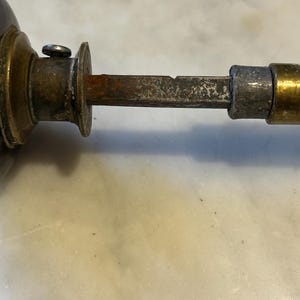May include: Close-up of an antique door handle. The handle features a dark brown knob, a brass collar, and a rusty, rectangular spindle. The metal components show signs of age and wear, with a tarnished appearance.