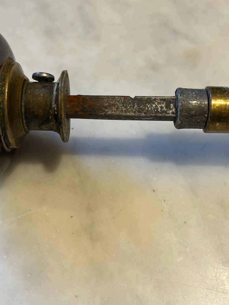 May include: Close-up of an antique door handle. The handle features a dark brown knob, a brass collar, and a rusty, rectangular spindle. The metal components show signs of age and wear, with a tarnished appearance.