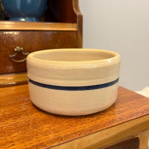 May include: A small, round stoneware bowl with a cream-colored exterior and a single navy blue stripe. The bowl is sitting on a wooden surface. A blue vase is visible in the background.