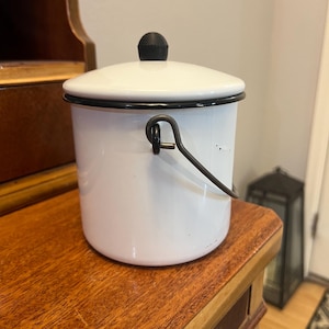 May include: A white enamel bucket with a black rim and handle. The bucket has a lid with a black knob. The bucket is sitting on a wooden surface.