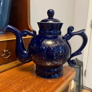 May include: A navy blue ceramic teapot with a gold design of buildings and text. The teapot has a rounded body, a curved spout, and a handle with a decorative swirl. The lid has a small knob. The teapot is sitting on a wooden surface.