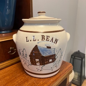 May include: Cream-colored ceramic jar with a lid, featuring the text "L.L. BEAN" and a hand-painted cabin scene in brown and blue. The jar has a brown rim and handles, and sits on a wooden surface.