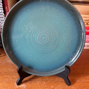 May include: A teal-colored ceramic plate with a spiral design in the center. The plate has a dark rim and is displayed on a black wooden stand. The plate is likely for serving food or as a decorative item.