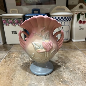 May include: A vintage ceramic vase in soft pastel colors. The vase features a light pink ruffled rim, handles, and floral relief details. The base is a light blue color. A decorative piece for home decor.