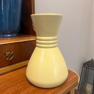 May include: A pale yellow ceramic vase with a bulbous base and a flared top. The vase has three horizontal ridges around the neck. A blue vase is visible in the background. The vase is sitting on a wooden surface.