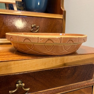 May include: A terracotta-colored, shallow ceramic bowl with a decorative pattern of intersecting arches and lines. The bowl is set on a wooden surface, showcasing its warm, earthy tones. The bowl's rim has a textured design, adding visual interest to this kitchenware item.