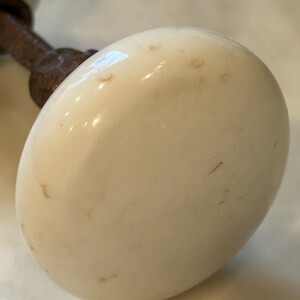 May include: A round, off-white ceramic door knob with a smooth, slightly weathered surface featuring small brown spots. The knob is attached to a dark, textured, rusted metal spindle that extends outward from the back of the ceramic piece.