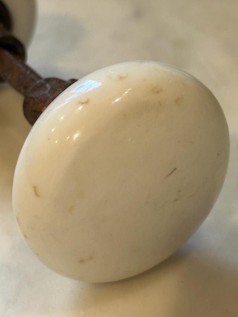 May include: A round, off-white ceramic door knob with a smooth, slightly weathered surface featuring small brown spots. The knob is attached to a dark, textured, rusted metal spindle that extends outward from the back of the ceramic piece.