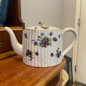 May include: White porcelain teapot with a lid, decorated with purple floral patterns and gold trim. The teapot has a curved spout and handle. The design includes vertical stripes and a gold-colored knob on the lid. A vintage tea set item.