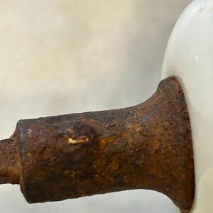 May include: A white ceramic knob attached to a cylindrical metal base with a heavily rusted, textured, and pitted surface. The metal component shows significant oxidation and corrosion, contrasting with the smooth, glossy white finish of the knob.
