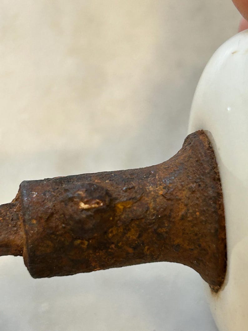 May include: A white ceramic knob attached to a cylindrical metal base with a heavily rusted, textured, and pitted surface. The metal component shows significant oxidation and corrosion, contrasting with the smooth, glossy white finish of the knob.