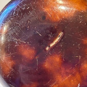 May include: Close-up of a round, dark brown object with a glossy finish. The surface has a mottled appearance with lighter brown and orange hues, and is marked with numerous fine scratches and a crack.