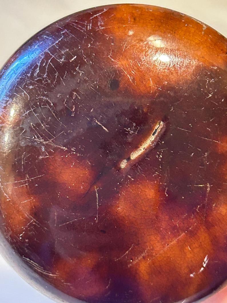 May include: Close-up of a round, dark brown object with a glossy finish. The surface has a mottled appearance with lighter brown and orange hues, and is marked with numerous fine scratches and a crack.