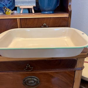May include: A rectangular cream-colored enamel baking pan with a thin teal rim. The pan has a smooth interior surface and a small hole on the upper right edge, designed for hanging or storage, showcasing a simple and functional kitchen design.