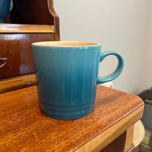 May include: A teal ceramic mug with a rounded handle. The mug has a light tan interior and is sitting on a wooden surface. The mug is approximately 4 inches tall.
