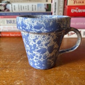 May include: A blue and white speckled ceramic mug with a curved handle. The mug has a wide rim and a cylindrical shape. The background includes a stack of cookbooks on a wooden surface.