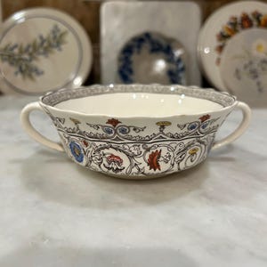 May include: Cream-colored soup bowl with two handles, featuring a floral and scroll design in blue, red, and gold. The rim has a decorative border. The bowl is on a white surface.