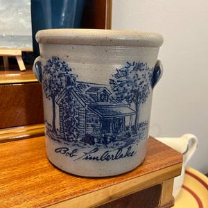 May include: A cylindrical stoneware crock with a speckled light gray finish and two side handles. The front features a blue embossed illustration of a cabin nestled between two trees, with the signature Bob Timberlake printed in blue script below.