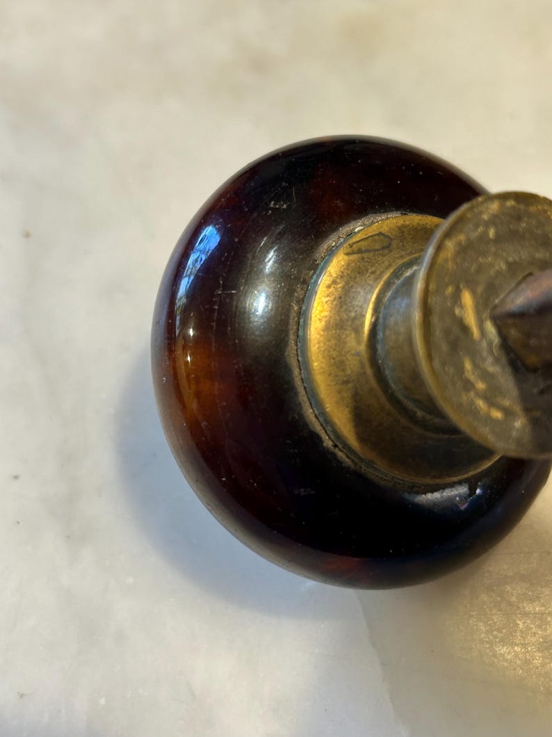 May include: Close-up of a vintage door knob. The knob is a dark, glossy brown with a brass base and a circular brass plate. The knob has a rounded shape and a smooth, reflective surface.