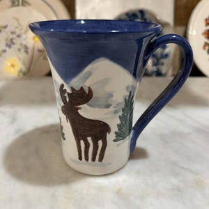 May include: A ceramic mug with a blue rim and handle, featuring a hand-painted design. The design includes a brown moose, evergreen trees, and a snowy mountain scene. The mug has a white base.
