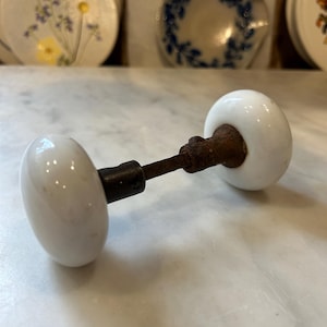 May include: A vintage door knob set featuring two smooth, round, white porcelain knobs connected by a central metal spindle. The metal components exhibit visible signs of oxidation and rust, contrasting with the clean, glossy white finish of the ceramic knobs, which are positioned horizontally on a flat surface.