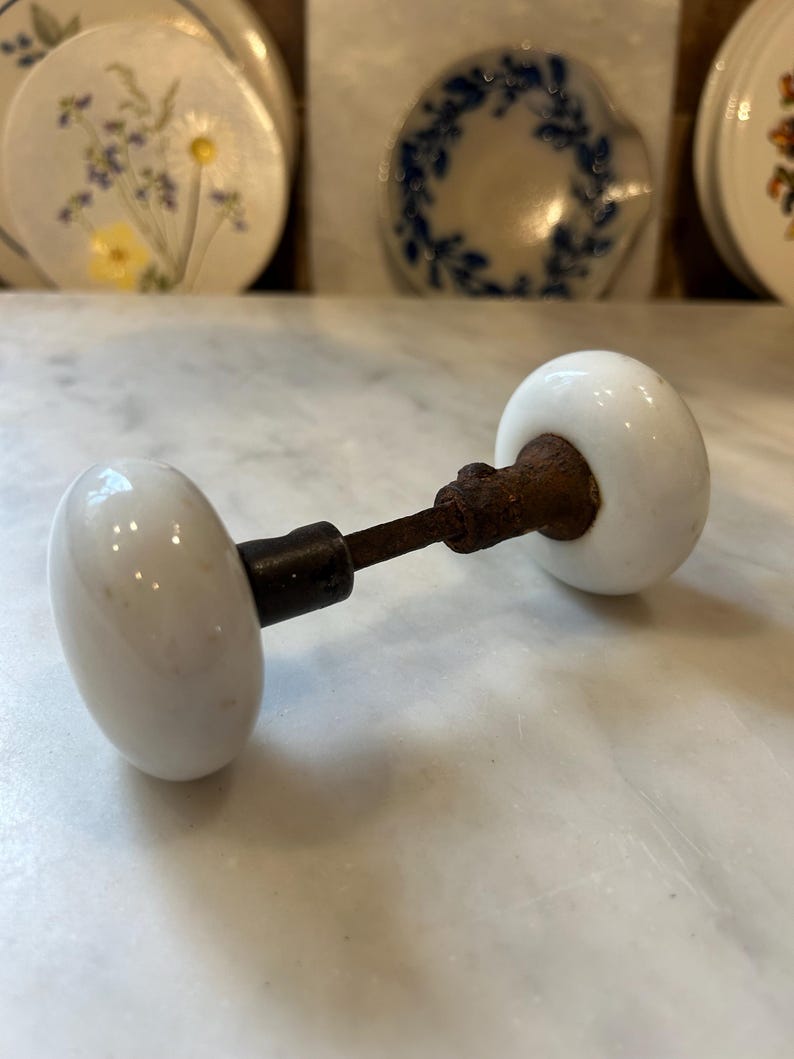 May include: A vintage door knob set featuring two smooth, round, white porcelain knobs connected by a central metal spindle. The metal components exhibit visible signs of oxidation and rust, contrasting with the clean, glossy white finish of the ceramic knobs, which are positioned horizontally on a flat surface.