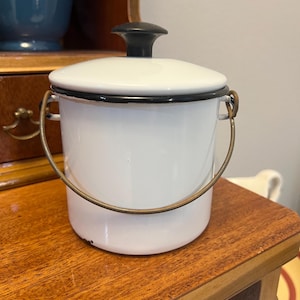 May include: A white enamel canister with a black rim and a matching white lid featuring a black knob. The container includes a thin, curved metal wire handle attached to two side loops, providing a simple and functional design for storage.