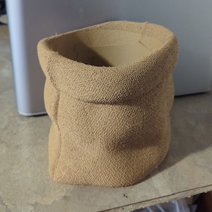 May include: A brown, textured, 3D printed planter shaped like a burlap sack. The planter is sitting on a wooden surface. The text "Bambu Lab" is visible on a white background in the upper right corner of the image.