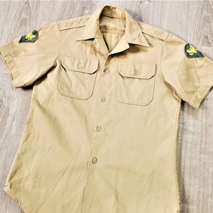 以下が含まれることがあります： ベージュの半袖シャツで、ポケットが2つとボタンが付いています。シャツの胸には黄色と緑のワッペンが2つ付いています。