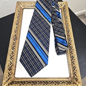 May include: A navy blue tie with a grid pattern and blue and gold stripes is draped over a gold framed mirror. The tie is made of a smooth fabric and has a classic design. The mirror is rectangular and has an ornate gold frame.