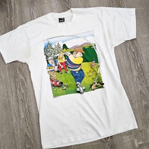 May include: White t-shirt with a cartoon illustration of three golfers on a green. The golfer in the foreground is swinging a golf club. The text "© 1988 C.C.A." is printed at the bottom of the illustration.