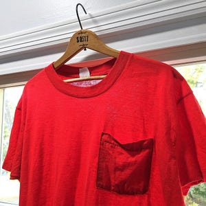 May include: A vibrant red t-shirt with a matching pocket on the front. The shirt is hanging on a wooden hanger with the text "S0517" printed on it. The shirt has a classic crew neck design.