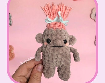 May include: A handmade crochet troll doll with a pink and gray color scheme. The doll has a textured body, small arms and legs, and a pink yarn hairstyle tied with blue ribbons. The text "Mildred Troll" and "No-Sew Crochet Pattern" are displayed.