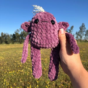 May include: A purple crocheted plush toy with a white yarn hair tuft and black button eyes. The toy has long arms and legs and is being held in a hand against a background of a field of yellow flowers.