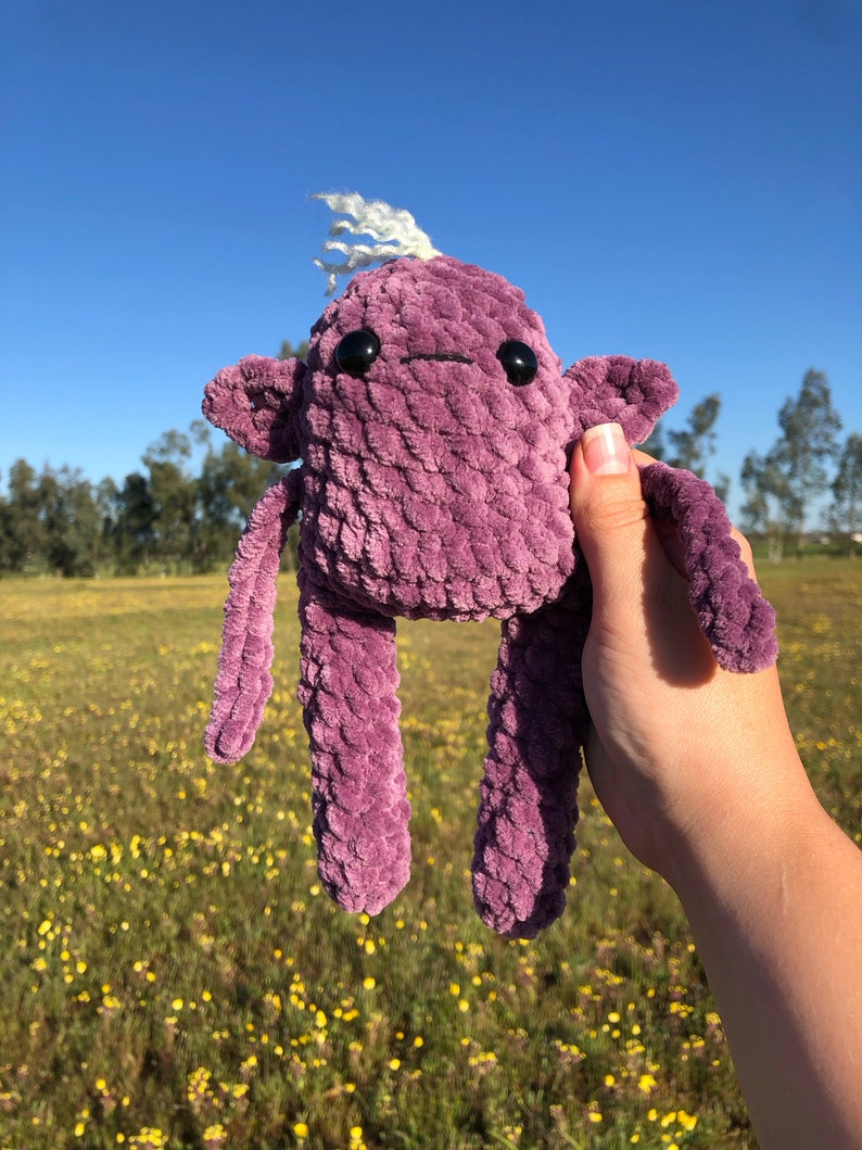 May include: A purple crocheted plush toy with a white yarn hair tuft and black button eyes. The toy has long arms and legs and is being held in a hand against a background of a field of yellow flowers.
