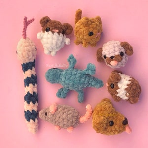Puede incluir: Una colección de peluches de animales de crochet en varios colores, incluyendo un lagarto azul, un gato marrón y blanco, un gusano rosa y azul, un ratón gris, un perro marrón y blanco, un hámster marrón y blanco y una oveja morada y blanca.
